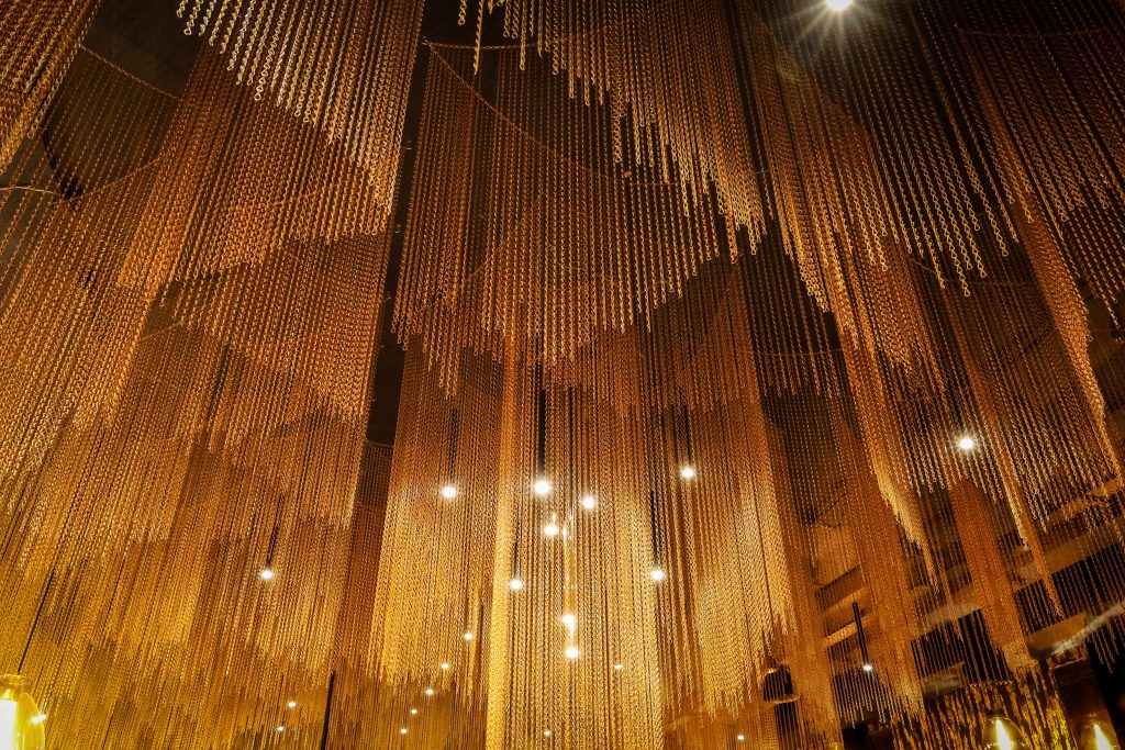 Ceiling installation of hanging golden bead strands lit by warm spotlights