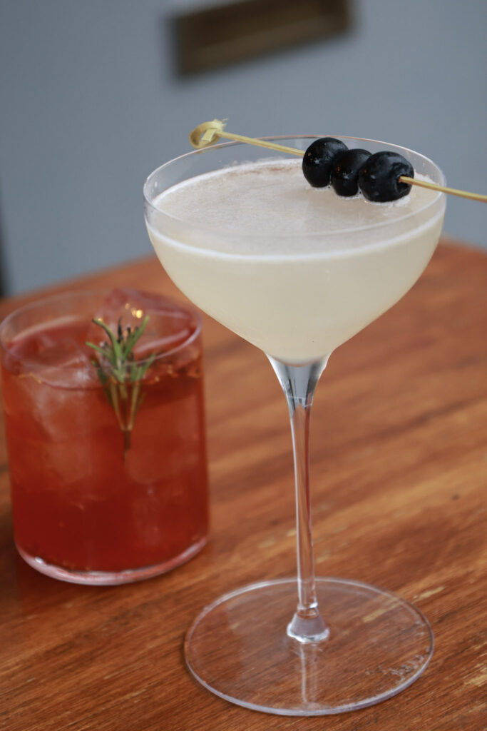 White cocktail with a blueberry garnish beside a red spritz