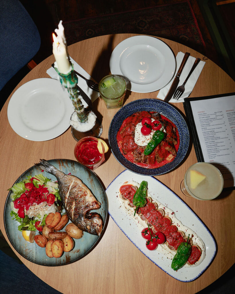 Overhead view of a shared table with grilled fish, small plates and cocktails