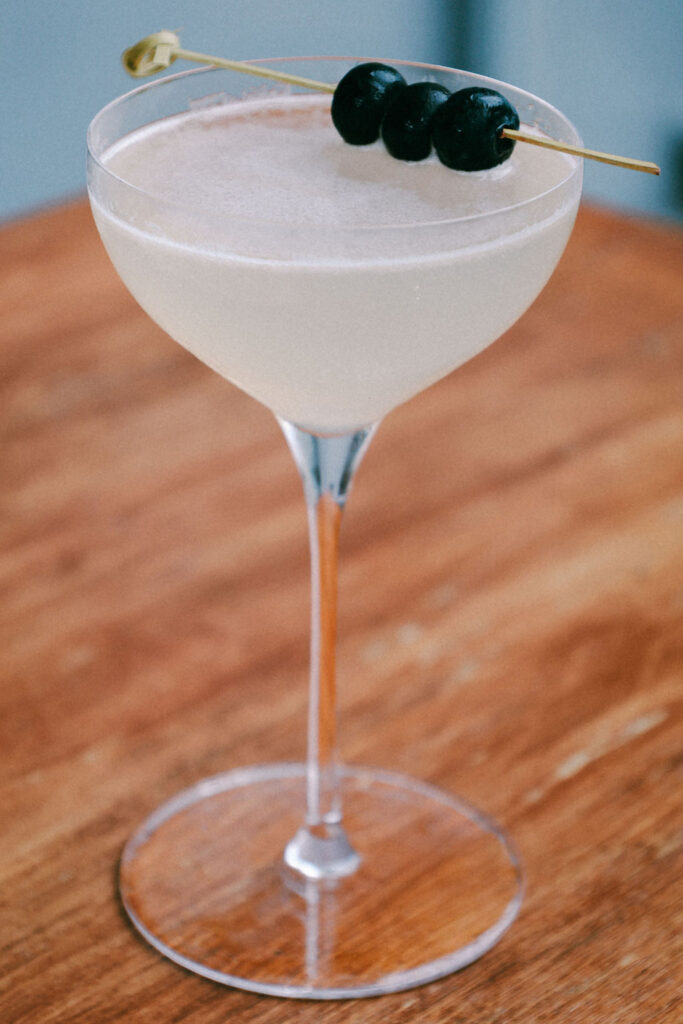 Pale cocktail in a coupe glass garnished with dark cherries on a wooden table